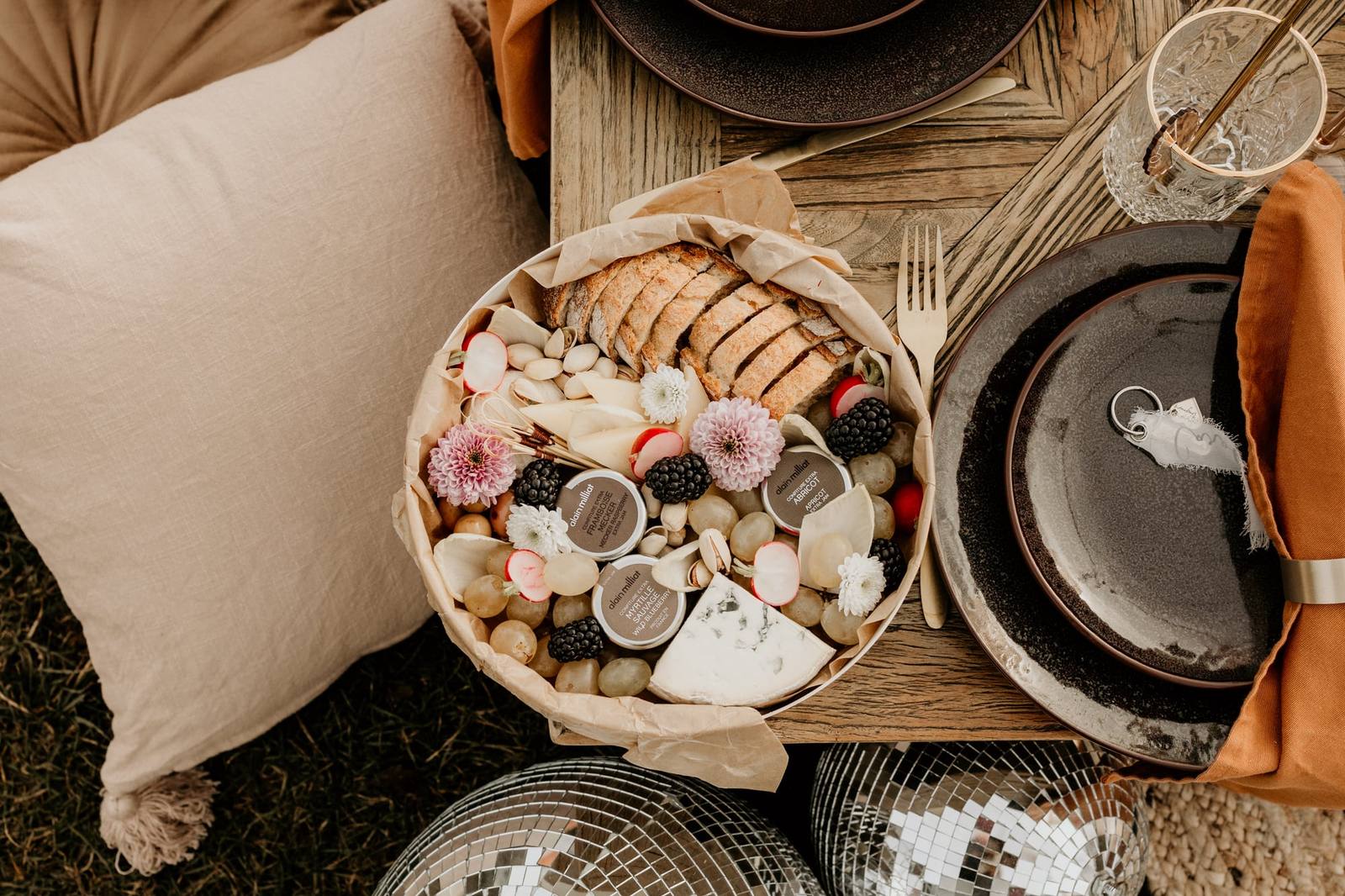 Box garnie de pain, fromages, raisins, mûres et fleurs comestibles sur une table dressée en plein air